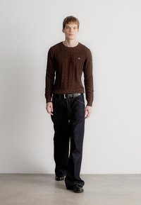 Καφέ πουλόβερ με πλέξη καλωδίου και μακριά μανίκια, συνδυασμένο με μαύρα παντελόνια τύπου wide-leg και μαύρα παπούτσια, σε ουδέτερο φόντο.