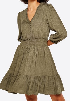 Nainen pitää yllään oliivinvihreää, teksturoitua mekkoa, jossa on 3/4-pituiset puhvimaiset hihat, napillinen V-aukko, smokkivyötärö ja röyhelöinen helma.