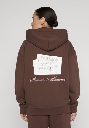 Frau mit dunklen Haaren zu einem Dutt gebunden, trägt einen braunen Hoodie mit weißem Grafikaufdruck und dem Text "Moments to Memories" auf der Rückseite, von hinten gesehen.