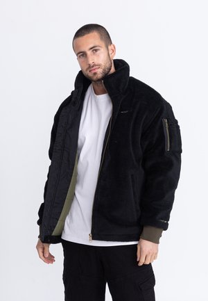 Mann mit kurzen Haaren, der eine schwarze Fleecejacke über einem weißen Hemd trägt, teilweise geöffnet, vor einem schlichten weißen Hintergrund stehend.