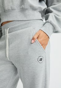 Pantalones de chándal de algodón gris con cierre de cordón, bolsillos laterales y un detalle de logo circular. Textura suave y corte relajado visible.