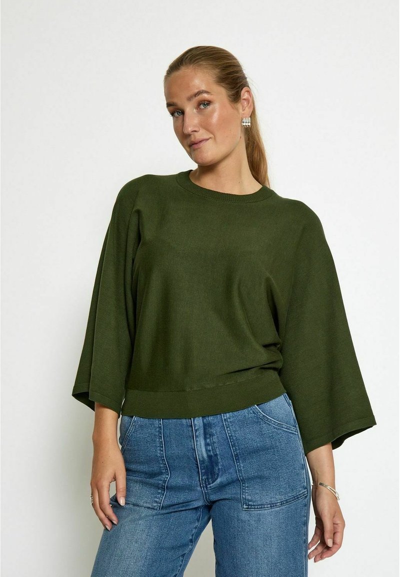 Pull à manches larges de couleur vert foncé, surdimensionné, avec un col rond, associé à un jean taille haute bleu clair. Texture en tricot simple.