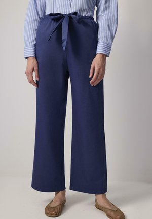 Pantalones de pierna ancha color marino con una textura suave, presentan una cinturilla elástica y un cinturón de tela atado. Combinados con zapatos planos trenzados.