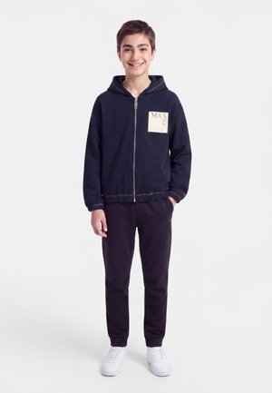 Garçon souriant debout, les mains dans les poches, portant un sweat à capuche zippé bleu marine avec écusson et pantalon assorti, baskets blanches, sur fond blanc uni.
