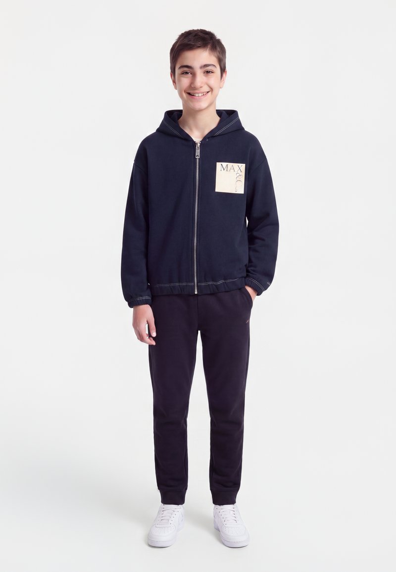 Garçon souriant debout, les mains dans les poches, portant un sweat à capuche zippé bleu marine avec écusson et pantalon assorti, baskets blanches, sur fond blanc uni.