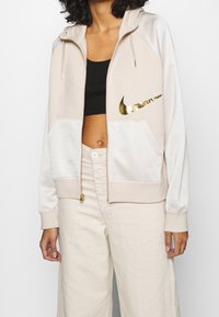 Beige zip-up hoodie med en guld Nike-swoosh. Har mjukt tyg, dragsko i huven och stora framfickor. Raglanärmar i off-white.