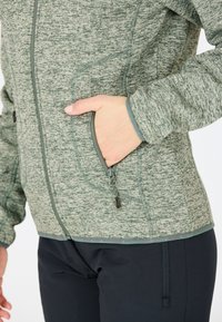 Person som bär en zippar, texturerad grön jacka med handen i fickan och svarta byxor där en sidoficka är synlig.