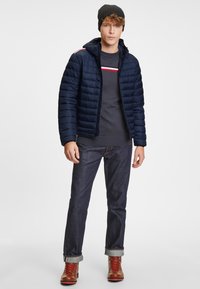 Marinblå puffjacka med horisontell sömnad, grå tröja med röda och vita ränder, mörka jeans och bruna snörade stövlar. Svart beanie.