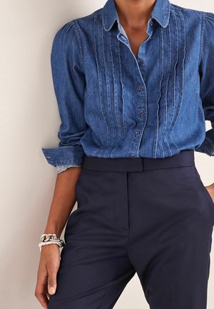 Denim-Hemd mit Knöpfen und Biesen-Details, kombiniert mit hoch taillierten marineblauen Hosen und silbernen Kettenarmbändern am linken Handgelenk.