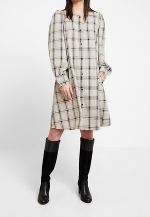 Femme portant une robe beige à carreaux boutonnée devant avec de longues manches bouffantes et des bottes noires montant jusqu'aux genoux, debout les mains dans les poches.