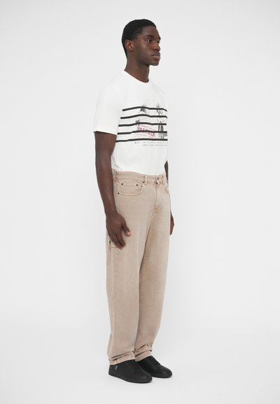 Άσπρο μπλουζάκι με οριζόντιες μαύρες και κόκκινες ρίγες, μπεζ άνετο τζιν παντελόνι και μαύρα αθλητικά παπούτσια. Απλός σχεδιασμός, χωρίς ορατά σχέδια ή λογότυπα.