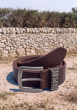 Un cinturón de cuero marrón oscuro con una gran hebilla metálica y el logotipo DIESEL grabado en relieve descansa sobre suelo seco delante de un muro de piedra.