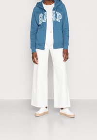 Felpa blu con zip e logo "GAP", t-shirt bianca, pantaloni bianchi a gamba larga e sneakers bianche con dettagli in paglia.