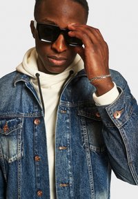 Denim jacket met koperen knopen, gedragen over een crèmekleurige hoodie. Model past zwarte rechthoekige zonnebril aan met een zilveren kettingarmband om de pols.