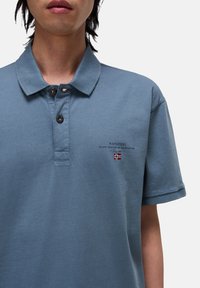Polo de color azul claro hecho de algodón, con un cuello de dos botones, logo y texto bordados, y un pequeño detalle de bandera en el pecho.