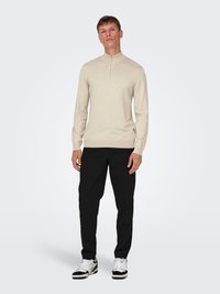 Cremfarbener Strickpullover mit Stehkragen und Reißverschluss, kombiniert mit schwarzen Hosen und schwarz-weißen Sneakers, vor einem neutralen Hintergrund stehend.