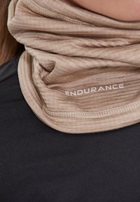 Beige halsduk av texturerat tyg, med en logo som säger "ENDURANCE" i vitt. Bärs över en svart topp.