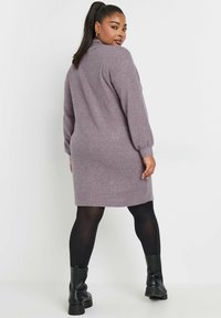 Langärmliges, lavendelfarbenes Kleid aus weichem, strukturiertem Stoff, mit hohem Kragen und leicht ausgestellter Form, kombiniert mit schwarzen Strumpfhosen und klobigen Stiefeln.