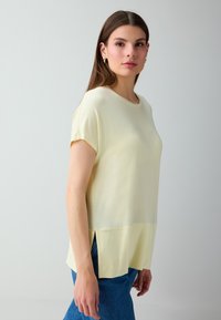 Jeune femme aux longs cheveux bruns, portant un haut jaune à manches courtes et un jean bleu, debout devant un fond gris clair uni.