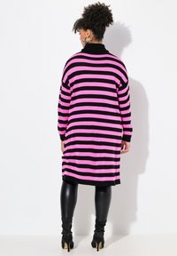 Pitkä neule, jossa on rohkeat vaaleanpunaiset ja mustat raidat, ribbireuna ja mustat yksityiskohdat. Käytetään mustien legginsien ja korollisten nilkkureiden kanssa.