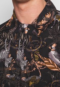 Zwarte button-up shirt met kleurrijke gedetailleerde prints van zebra's, vogels, slangen en bladeren, gedragen door een persoon die van de nek omlaag wordt getoond.