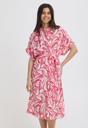 Mosolygó nő, rózsaszín és fehér levélmintás, derékban megkötött midi ruhában, rövid ujjú, egyszínű háttér előtt áll.