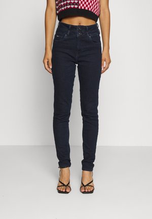 Dark blue skinny jeans feature a high waist with two buttons, five pockets, and a smooth denim texture. Worn with a cropped patterned top.