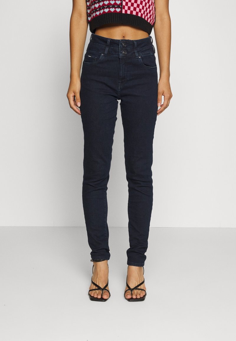 Los jeans skinny azul oscuro tienen una cintura alta con dos botones, cinco bolsillos y una textura de denim suave. Se combinan con una blusa corta estampada.