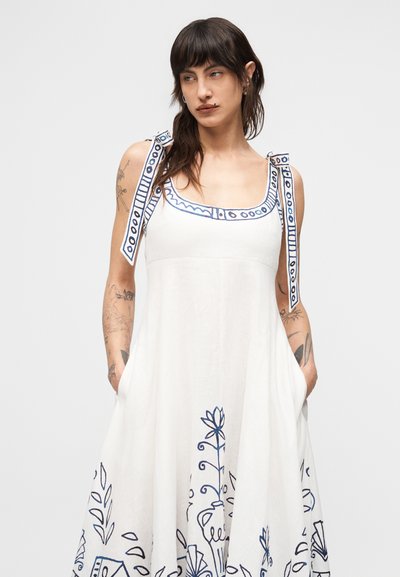 Γυναίκα με σκούρα μαλλιά που φορά άσπρο αμάνικο φόρεμα με μπλε λουλουδένια και γεωμετρικά σχέδια, χέρια στις τσέπες, με τατουάζ και στα δύο χέρια.