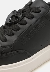 Melnas ādas sporta apavi ar teksturētu virsmu, aprīkoti ar melnām šņorēm un sirds formas akcentu. Sānos ir izcelta „LOVE MOSCHINO” markēšana.