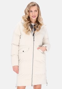 Vrouw in een lange, crèmekleurige gewatteerde parka met capuchon, voorzijde met rits en klepzakken, staand met één hand in de zak.