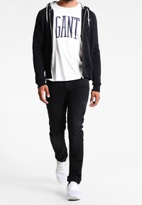 Felpa nera con zip sopra una maglietta bianca con la scritta "GANT" stampata in blu navy. Jeans neri e sneakers bianche completano l'outfit.