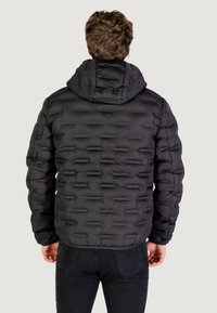 Giacca a vento nera con un design trapuntato e testurizzato, dotata di cappuccio e silhouette aderente. Il tessuto appare liscio e leggero.