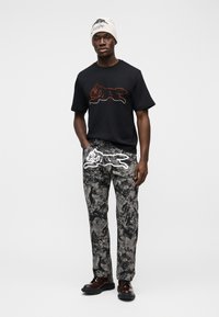 Μαύρο μπλουζάκι με σχέδιο κόκκινων στρας σε σχήμα γάτας, συνδυασμένο με γκρι παντελόνι με σχέδια και λευκό γραφικό γάτας. Το σύνολο ολοκληρώνουν καφέ παπούτσια.