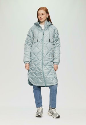 Veste longue matelassée bleu clair avec capuche, dotée d'une fermeture éclair intégrale, de cordons de serrage et d'un motif en diamant. Portée sur un jean et des baskets.