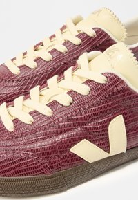 Αθλητικά παπούτσια χαμηλού προφίλ με υφασμένα μπορντό επάνω μέρη, εκρού κορδόνια, εκρού λογότυπο στο πλάι και καφέ λαστιχένιες σόλες.