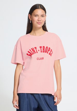 Jeune femme aux longs cheveux foncés portant un t-shirt rose avec "SAINT-TROPEZ CLUB" en lettres rouges et un pantalon bleu, debout sur fond blanc.