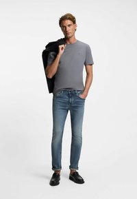 Camiseta gris ajustada con un pequeño logo, combinada con jeans ajustados de color azul claro y mocasines negros. El modelo lleva una chaqueta negra sobre el hombro.