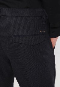 Marinblå mönstrade byxor med en strukturerad tyg; har en bakficka med en mörk trim och en liten orange detalj nära fickan.