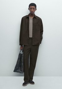Brown button-up shirt, beige top, and matching trousers; model holds a dark tote bag. Smooth texture, relaxed fit, minimal design.