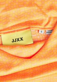 Jasnopomarańczowa tkanina z wzorem w kratkę. Metka pokazuje „JJXX” i rozmiar „S”. Wyprodukowane w Bangladeszu. Gładka, lekka textura.