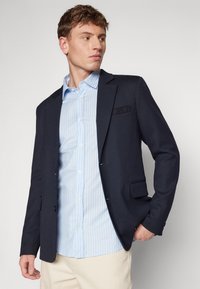 Navy blazer with notch lapels, single chest pocket, and two front pockets, worn over a light blue striped shirt, paired with beige pants.