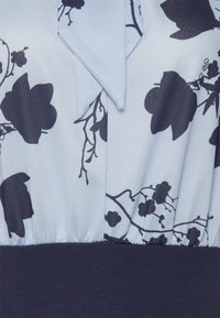Blouse bleu clair avec des motifs floraux sombres, un col, et une taille froncée avec une bande unie marine en bas.