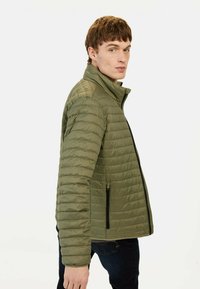Olivengrøn quiltet jakke med ståkrave, lynlåslukning foran og to side lommer, der har et slankt, polstret design og struktureret overflade.