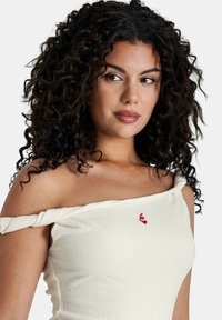 Haut crème à épaules dénudées en tissu texturé, avec un détail rouge brodé et des manches nouées, offrant une silhouette ajustée.