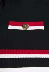 Schwarzes Strickkleidungsstück mit einem dünnen roten und weißen Streifen. Enthält ein dekoratives goldenes Emblem mit verschlungenen Schlaufen auf einer oberen Tasche.