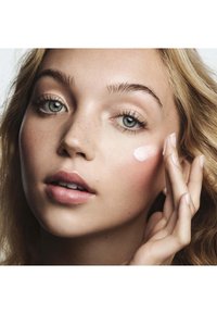 Model with light skin and tousled blonde hair applying a creamy pink product to her cheek; visible freckles and soft, natural makeup finish.
