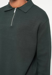 Mørkegrønn, strikket genser med krage og halv-glidelås. Har et teksturert stoff med en tettsittende design og ribbestrikkede mansjetter.