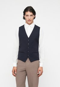 Colete azul-marinho com seis botões, lapelas pontiagudas e dois bolsos frontais, usado sobre uma camisa branca de manga longa e calças bejes.