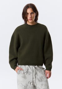 Pull en maille vert olive avec col rond, épaules tombantes et ourlet côtelé. Assorti à un pantalon gris clair avec des motifs abstraits.
